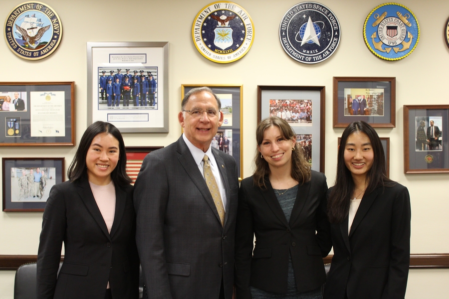 Photo of Michelle Lin, Senator John Boozman, Alyssa Rudelis, and Diane Zhang posing for a photo in front of a wall with many framed photos and service plaques 