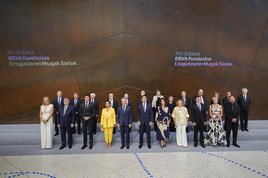 Twenty-four people in formal attire stand in front of the side of a building made with modern rusted industrial metal. Text behind them in light reads "XIV Edizioa BBVA Fundazioa Ezagutzaren Mugak Sariak"