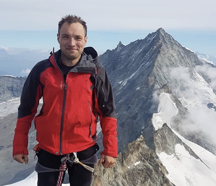 Photo of Octavian-Eugen Ganea, wearing a ski jacket and climbing ropes, standing high up on a mountain with another mountain peak behind him 