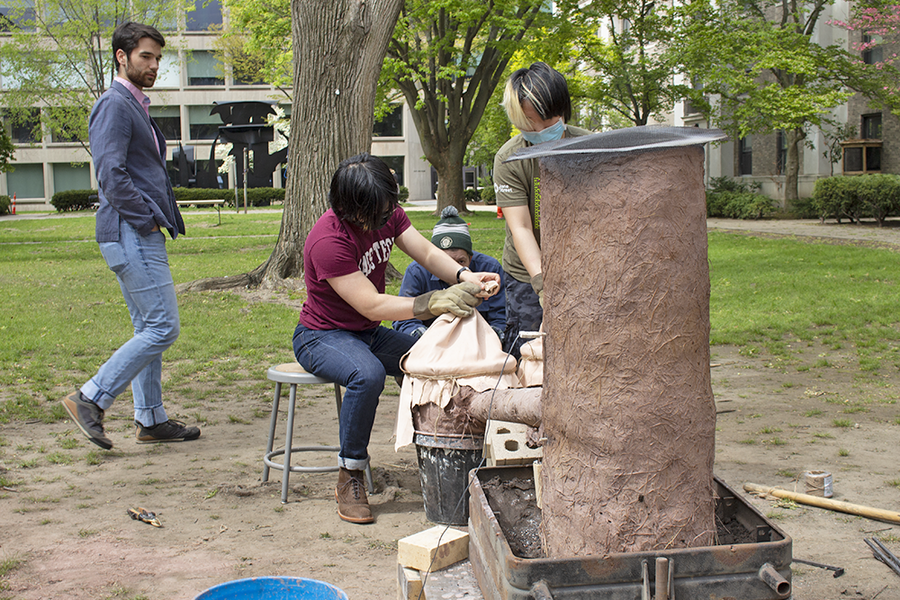 Photo of students working with a brown cylindrical furnace made from a mud-and-straw mixture that is nearly as tall as they are in a grassy courtyard