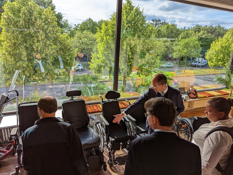 Photo of a man gesturing at a line of three unoccupied wheelchairs while three people observe. They are in a room with large windows loking out on trees.