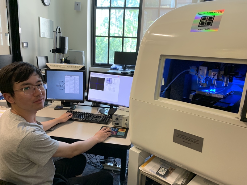 Photo of Jiahao Dong sitting at a desk with two monitors in Jaramillo lab. A large piece of lab equipment labeled “TriboIndenter” is in the foreground.