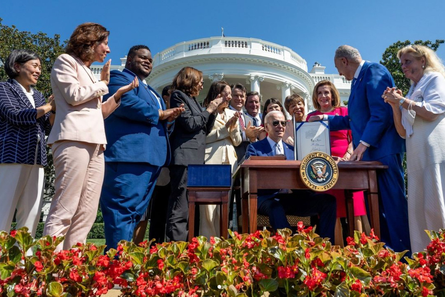 President Biden sits at a desk, while Senator Schumer stands nearby, propping up a signed document resting on the desk. A row of people stands behind the president, applauding, and the White House is in the background.
