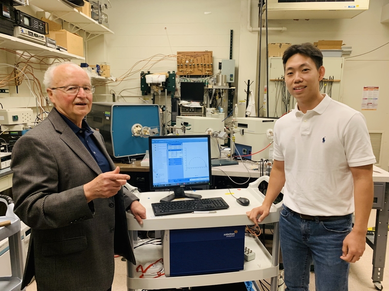 Harry Tuller and student pose for a photo in a lab, with a computer screen on a table between them showing data