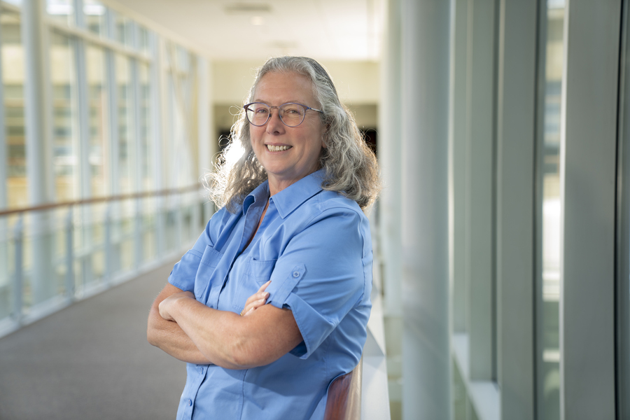Mary Ellen Zurko poses for photo, smiling in a sunlit hallway with her arms crossed in front of her.