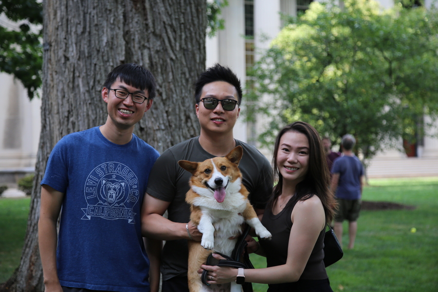 Three graduate students stand together in Killian Court. Two of them are holding a corgi dog who’s looking directly at the camera and seems to be smiling.