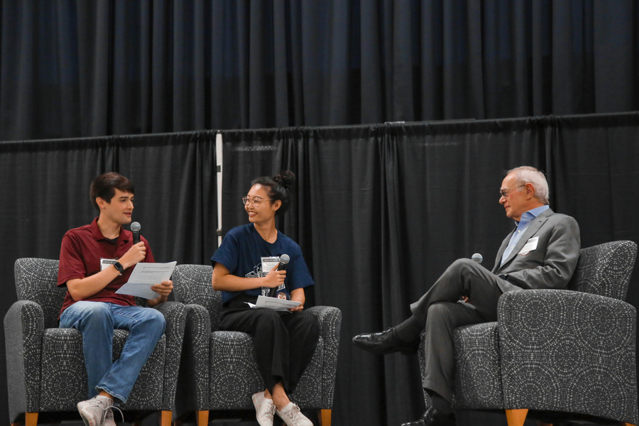 On a stage draped with black cloth, two grad student moderators and President Reif sit in comfortable chairs facing each other, holding microphones.
