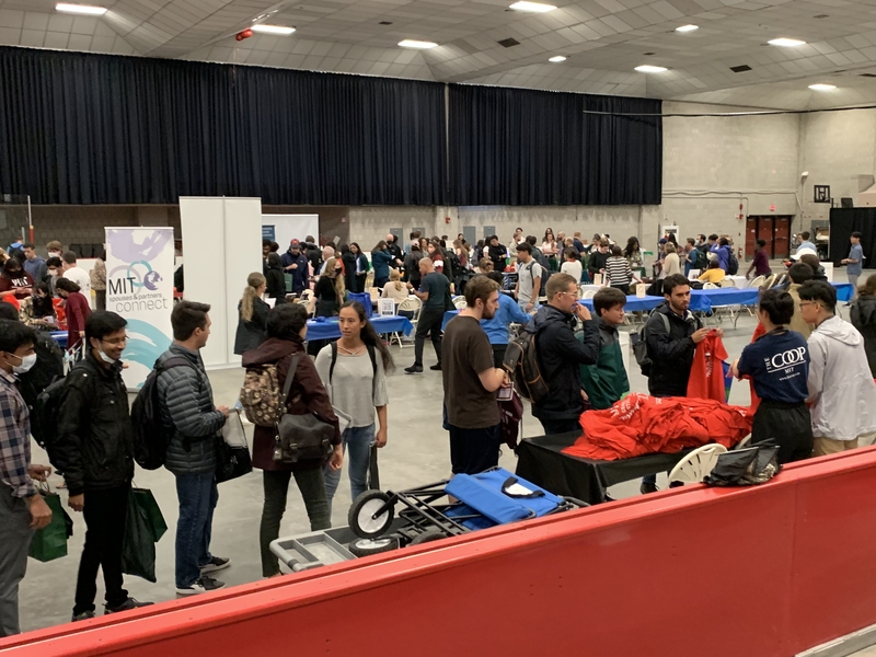 A line of graduate students wait to pick up brightly-colored T-shirts from a table at the Resource Fair. In the background, other tables and a crowd of students are visible.