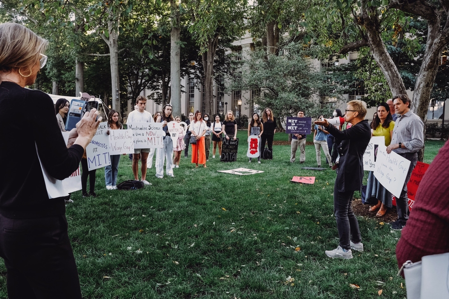 Photo of Linda Griffith with a bullhorn, addressing a semicircle of about 30 rally attendees on a grassy area, many holding hand-painted signs.