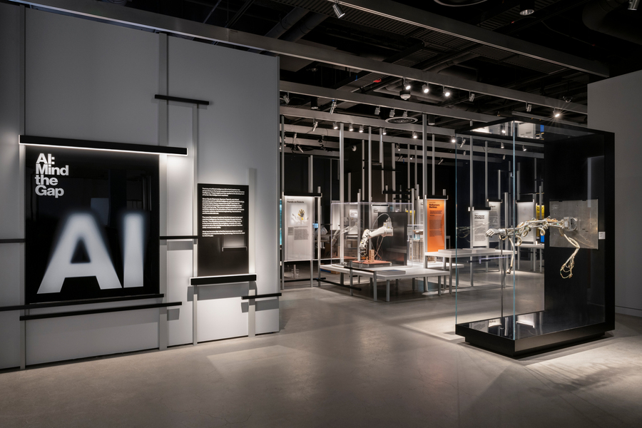 A black square sign says “AI: Mind the Gap,” with AI in giant letters. Numerous large glass-walled boxes hold robot arms of different types. 