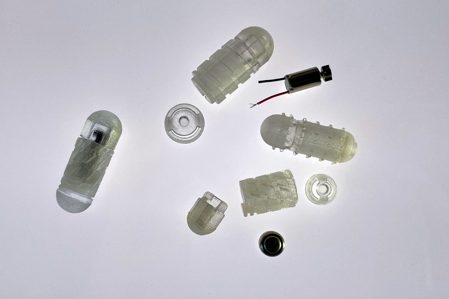 Nine pieces of the drug capsule prototype are on a white background. Some pieces are pill-shaped and made of translucent material. Some are threaded like a screw.  
