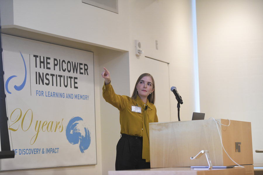 Indie Garwood stands at a podium during her presentation and points to her right (toward an unseen screen). Behind her is a sign showing the Picower Institute 20th Anniversary logo