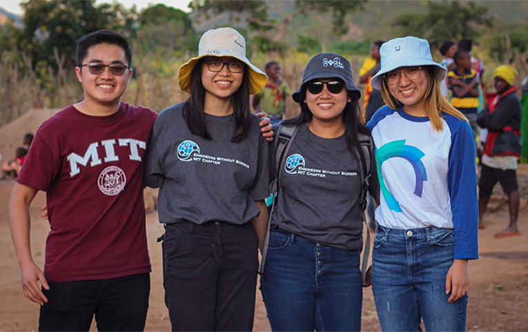 Four students wearing MIT T-shirts pose together with grasslands in the background. Half a dozen villagers of Mkutani, Tanzania, stand in the background.