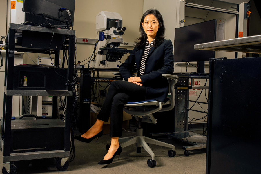 Gloria Choi sits on a chair surrounded by grey and black equipment in a lab, including a microscope, computer monitors, and rolling carts with devices on them. 