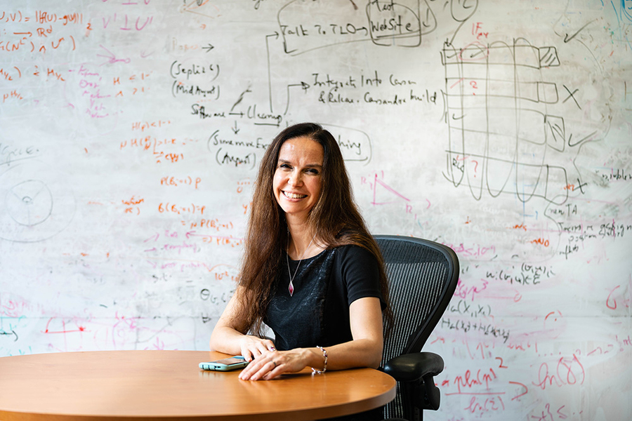 Portrait photo of Muriel Médard, seated in an office before a whiteboard with lots of equations written on it