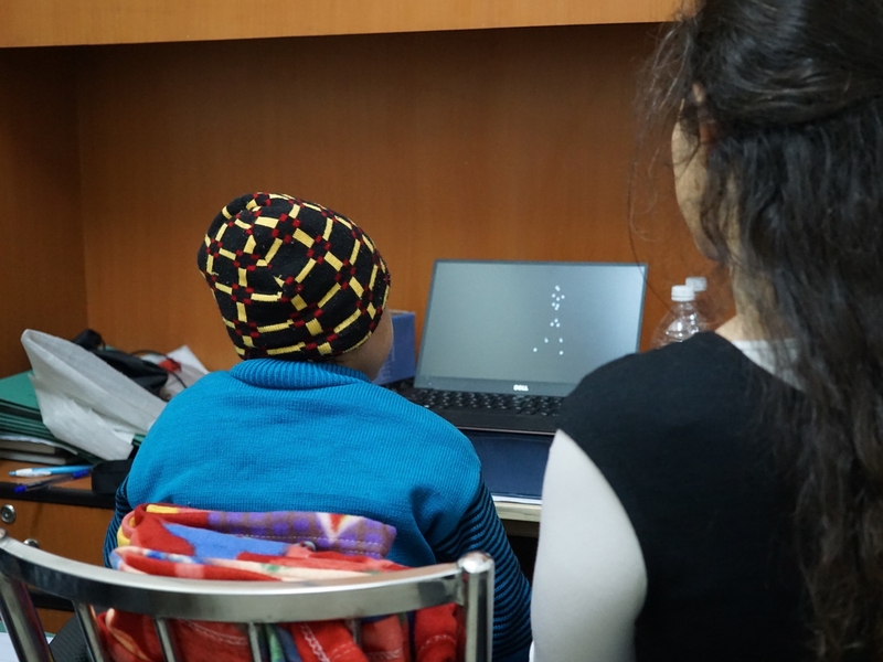 Photo from behind a person sitting at a desk, watching dot patterns on a monitor, while another person looks on. 