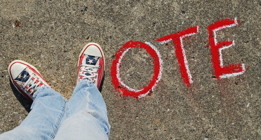 Top-down view of two feet wearing red, white, and blue sneakers positioned in the shape of a V next to letters written in chalk on the ground spelling the word "Vote"