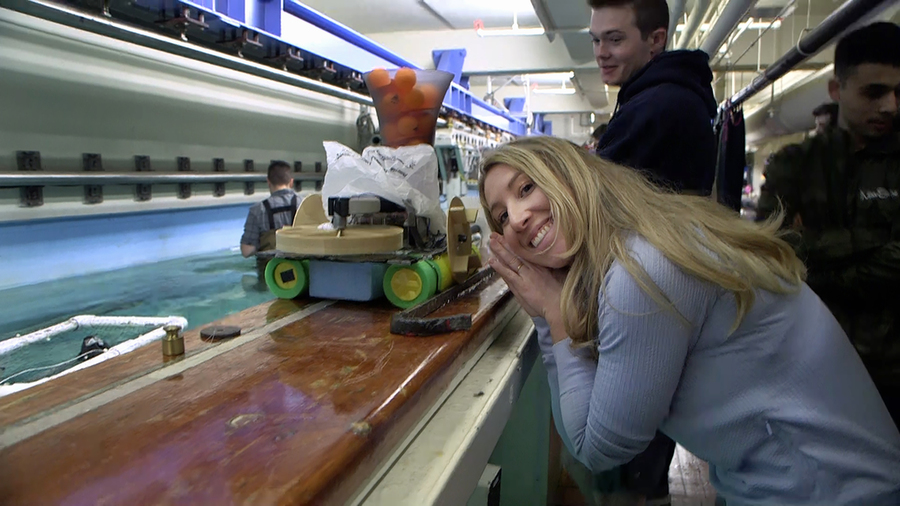 Woman leans on the ledge surrounding an indoor water tank, posing next to a small robotic boat that is also resting on the ledge