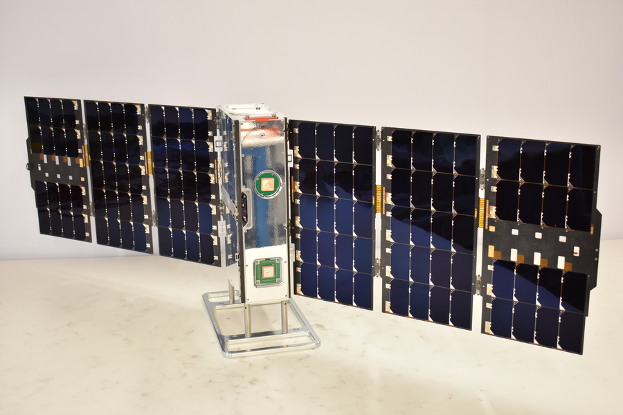 The Agile MicroSat, a tall, narrow, rectangular device, stands on a white surface with an array of panels spread out like wings on either side. Each array consists of three rectangular panels, each the same height and slightly wider than the MicroSat device, arranged side-by-side.