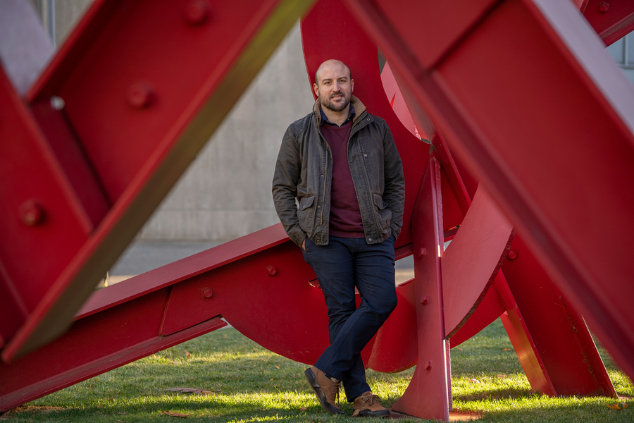 On a grassy courtyard, Otto Cordero stands inside a sculpture comprised of overlapping red steel beams