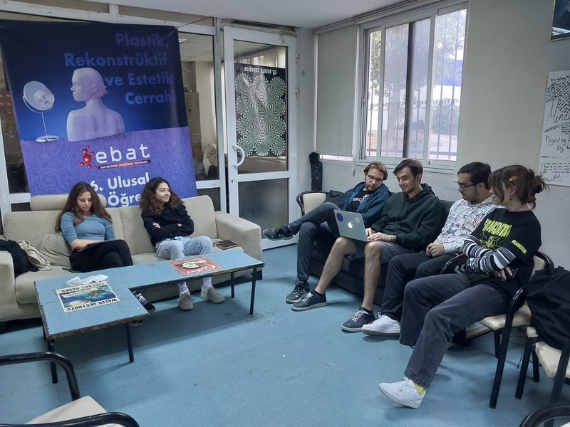 Photo of a medical student lounge with 2 couches and a small table. 2 students sit on the left couch with a poster of the school name behind them. 4 students on the right couch look at a laptop together.