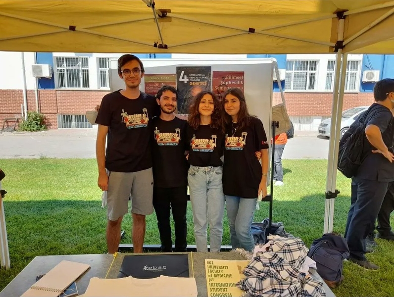 Photo of 4 medical students standing outside a school building underneath a tent, with informational posters behind them and flyers on the table in front of them.