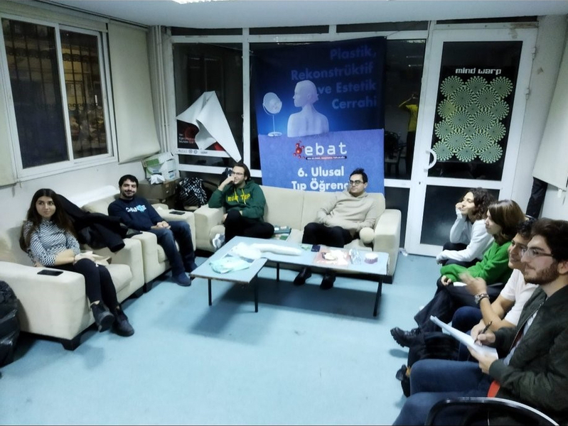 Photo of a student lounge with 8 students sitting in a semi-circle of couches and chairs, with a table between them. A poster with the school name is behind them.