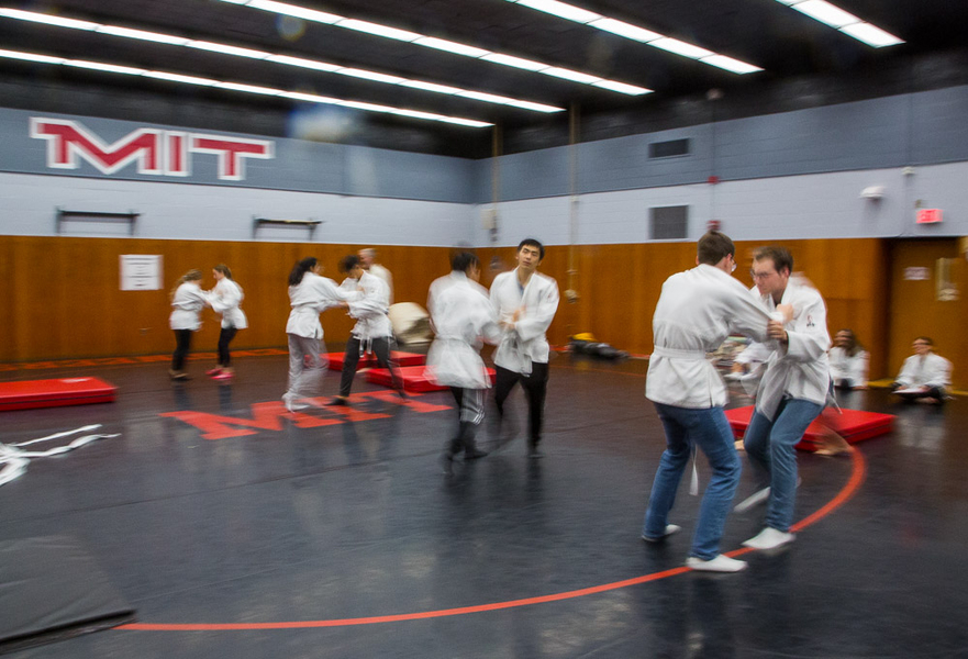 Students practice judo moves one-on-one in a wrestling gym where "M.I.T." is written on the wall