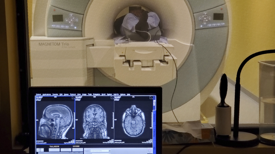 Control-room view of a person in an MRI machine. In the foreground on the bottom left is a monitor showing three views inside the person's brain.