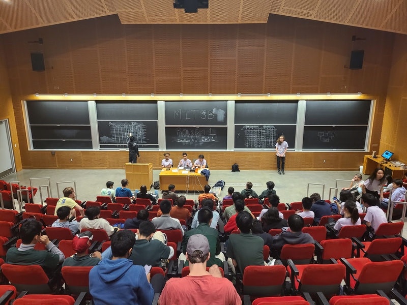 A semi-full lecture hall, photographed from the back of the room. Two teams of four high school students sit in the first row. Facing the audience in front of a large set of chalkboards are five volunteers in purple keeping score, operating the timer, and reading questions.