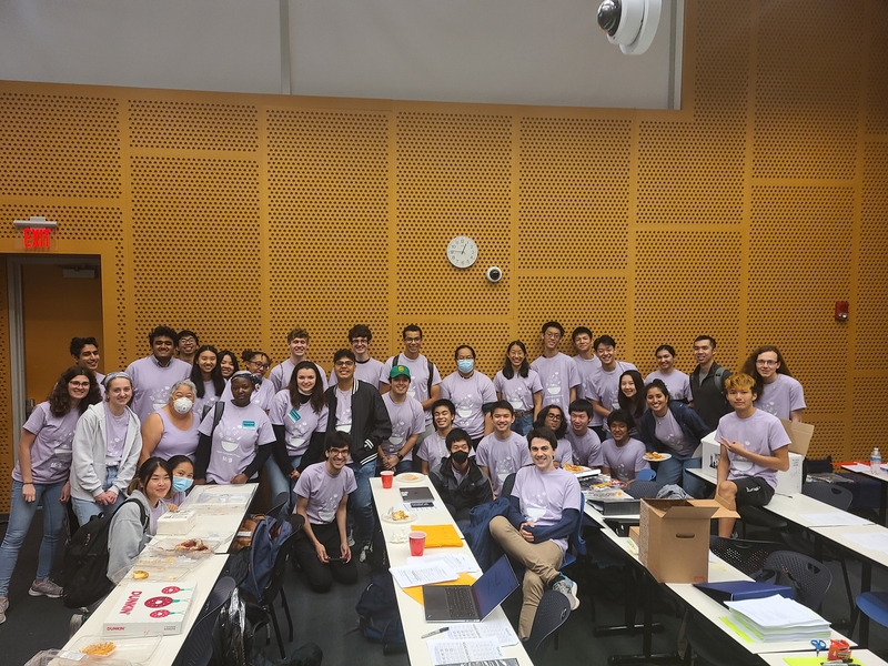 Science Bowl volunteers wearing purple shirts and posing together in a large group with tables in the foreground
