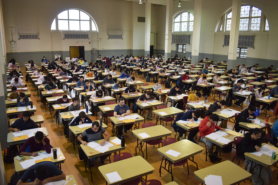 A large gymnasium-like room filled with students sitting at small desks, which are arranged in 12 rows