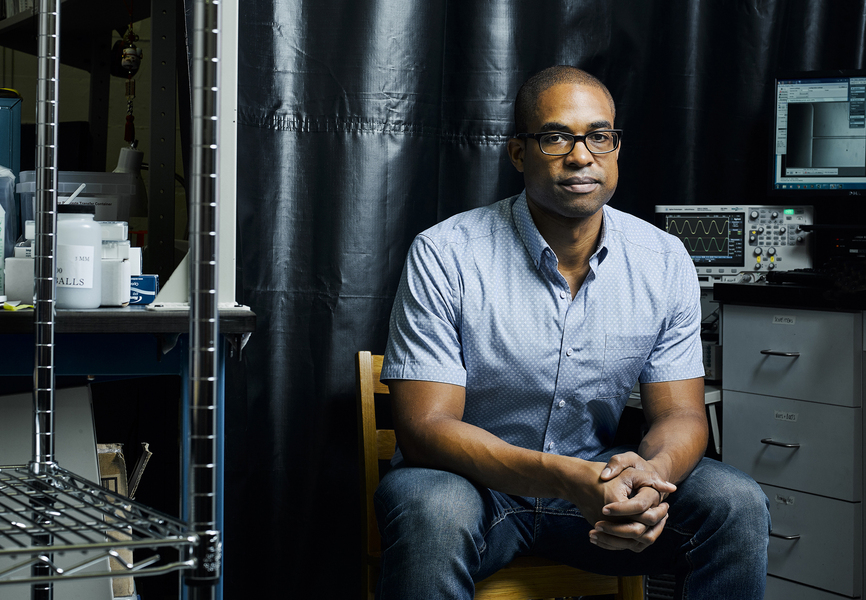 Photo of a man in a shortsleeve button-down shirt sitting in a lab with his hands folded, looking at the camera