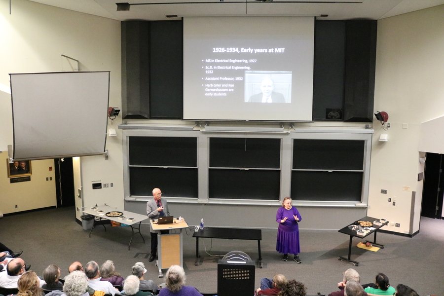 Photo of J. Kim Vandiver standing at a podium to the left of Edgerton Hall and an ASL interpreter to the right. Behind them, a slide is projected on the wall.