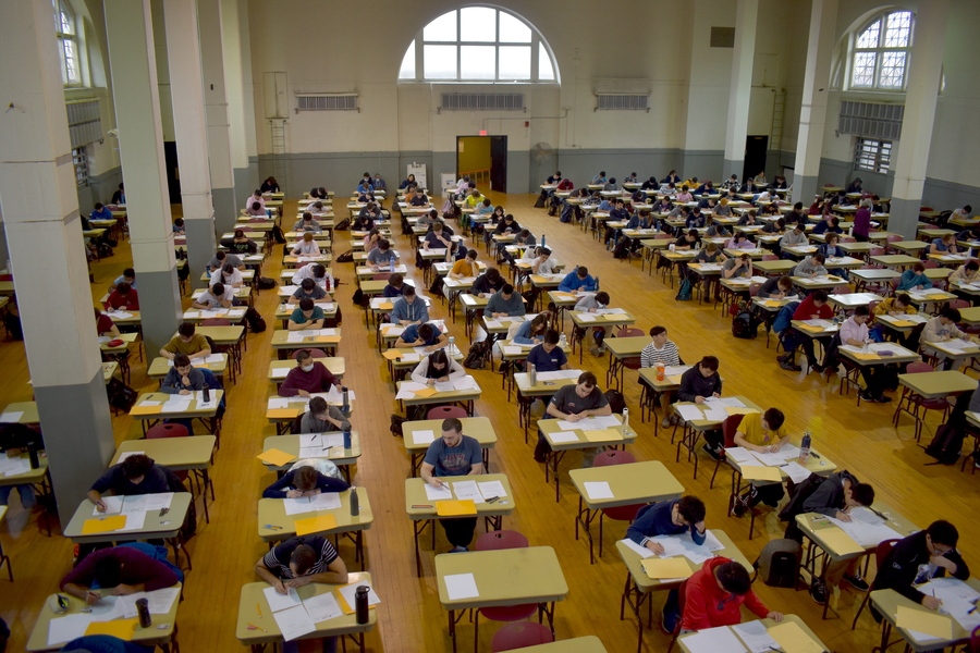A large room with at least 14 rows of about 20 identical desks, each with one student taking an exam.