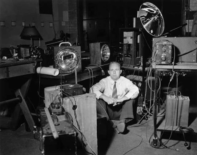 Historical black-and-white photo of Harold “Doc” Edgerton sitting on the floor in a room surrounded by strobe lights.