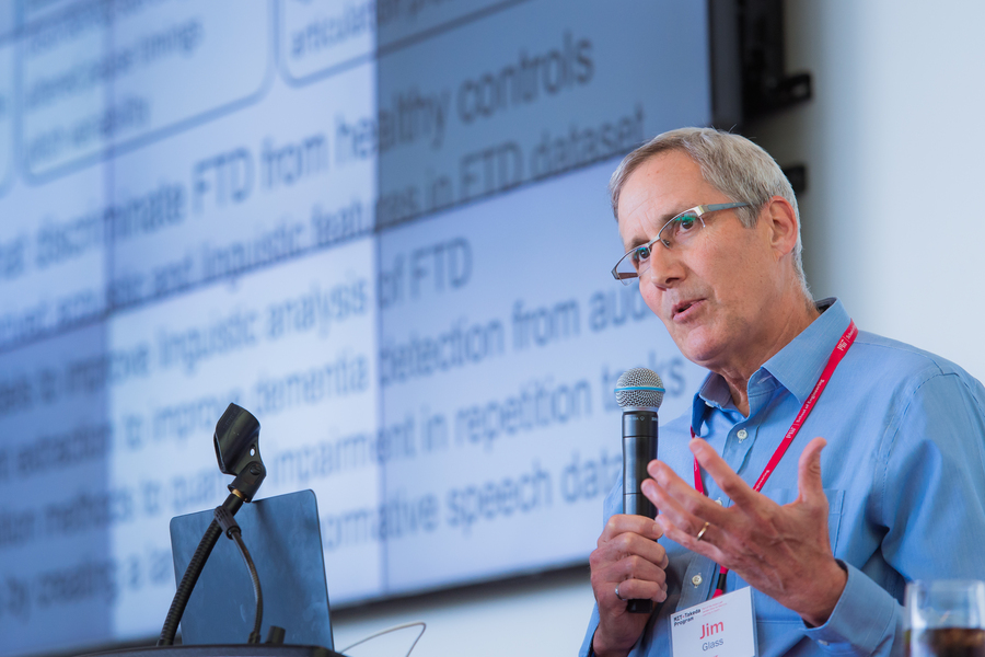 Jim Glass standing with a microphone in front of a projector screen at an MIT-Takeda Program event