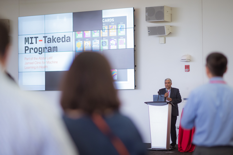 Camera focuses on Anantha Chandrakasan standing at a podium, as seen through the blurred backs of several audience members.