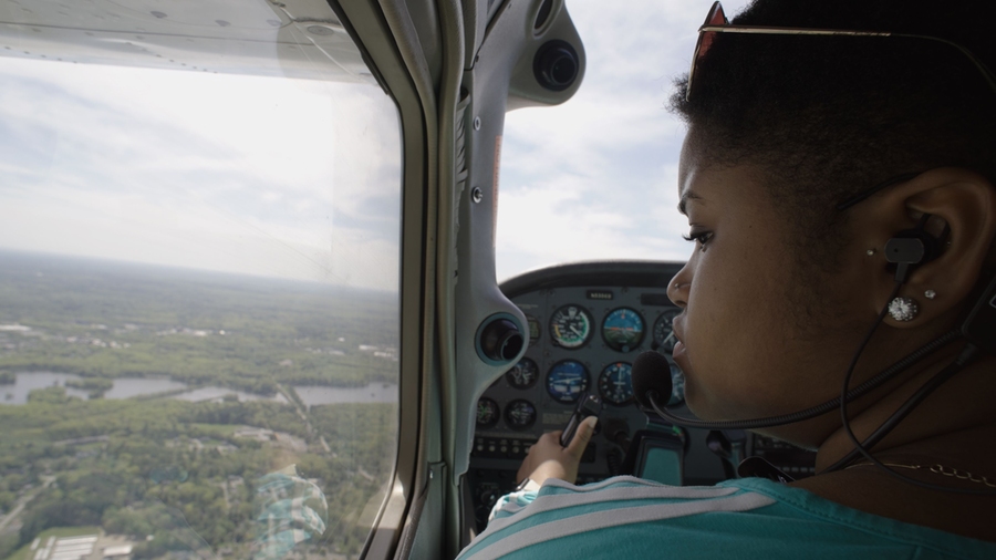 An over-the-shoulder close-up of Elissa Gibson flying a small plane. On the left is an aerial view of trees, houses, and a river. On the right, Gibson is looking out the window and steering the plane. She is wearing sunglasses on her head, a headset, and a turquoise shirt. The plane’s controls are visible over her shoulder.