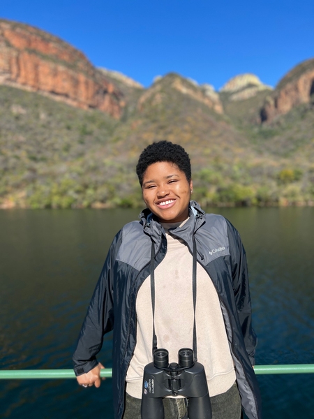 Elissa Gibson standing in front of a lake with mountains in the background. The sky is blue and cloudless. She is smiling and wearing binoculars around her neck.