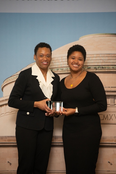 Elissa Gibson and Melissa Nobles hold the Bridge Builder Award between them. The women stand in front of a large indoor mural of the MIT dome.