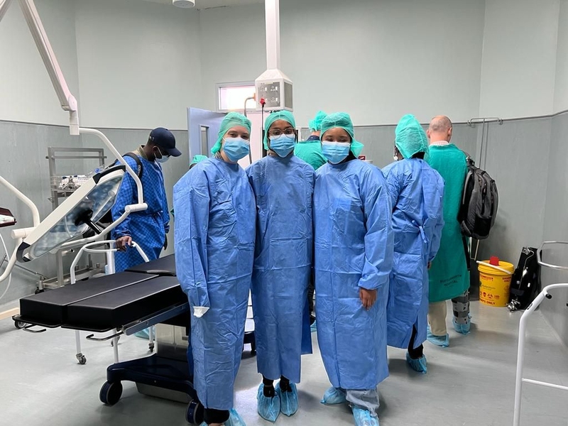 Three young adults are photographed in full scrubs — blue head covers, blue face masks, blue gowns, and blue shoe coverings — in a surgical room of a hospital. Elissa Gibson in on the right. Four other people can be seen in the background. The room is white, grey, and sterile looking. An empty black surgery table is behind the group.