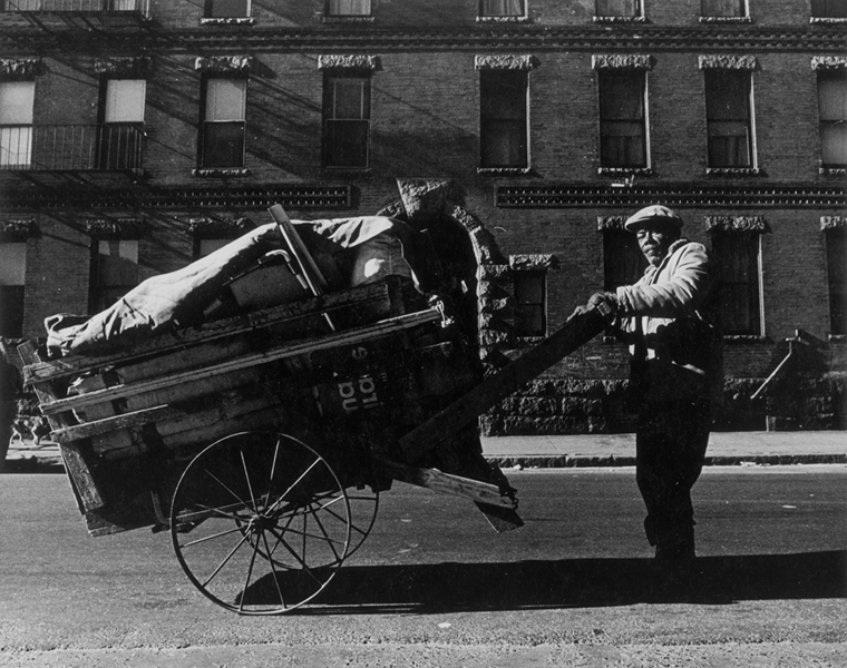 1960s black-and-white photo of a man pushing a two-wheeled cart on the street in front of a brick building