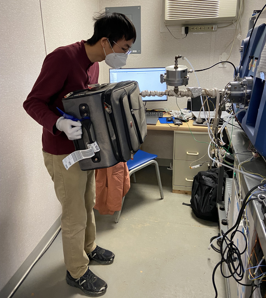 A man wearing a face mask and nitrile gloves holds a suitcase up to a mass spectrometer in a lab.