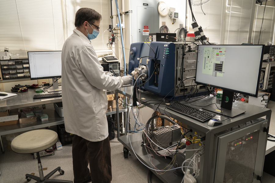 Ted Mendum stands in front of a mass spectrometer (an instrument a bit larger than a microwave, with wires protuding from it). Next to it is a computer screen showing data.