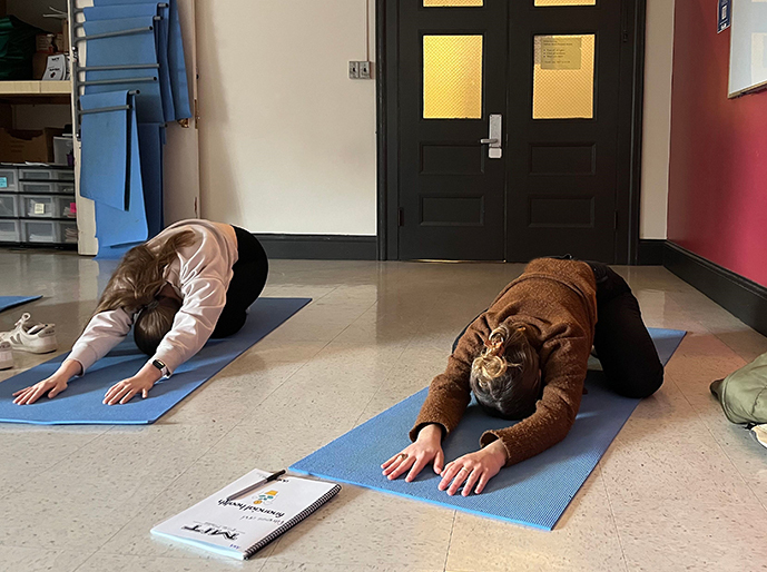 Two students on yoga mats kneel and stretch forward as far as they can, faces down and hands flat on the floor in front of them.