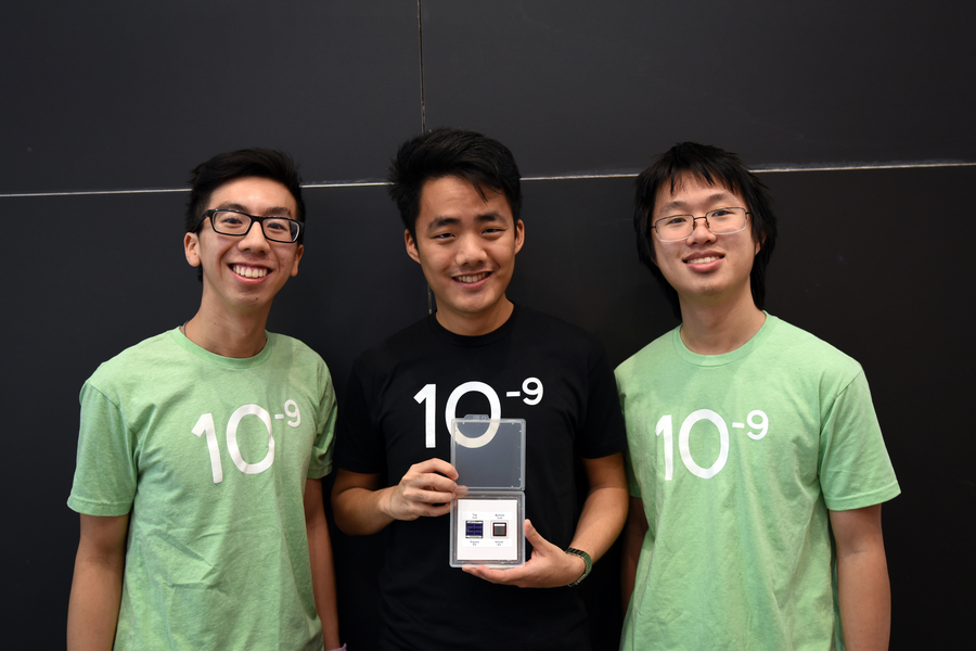 Three men wearing MIT.nano T-shirts reading "10 to -9" stand in front of a blackboard. The student in the center holds a plastic case displaying their photovoltaic device.