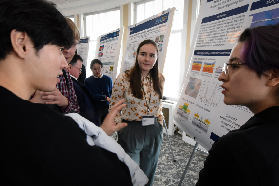 Flanked by two other students asking questions, a woman stands in front of a posterboard detailing the class project.