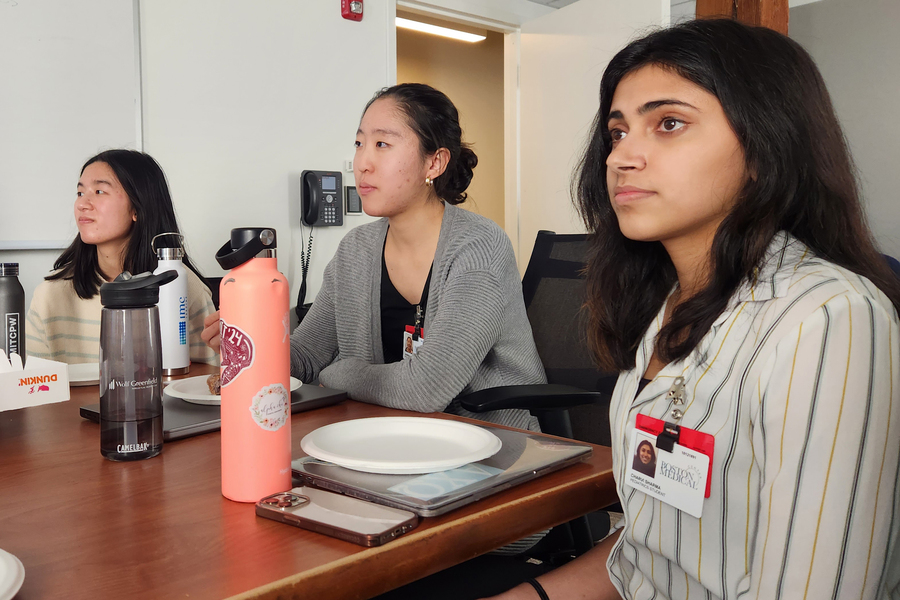 3 people, wearing Boston Medical Center badges, watch a presentation in a conference room, with drinks and snacks on the table.