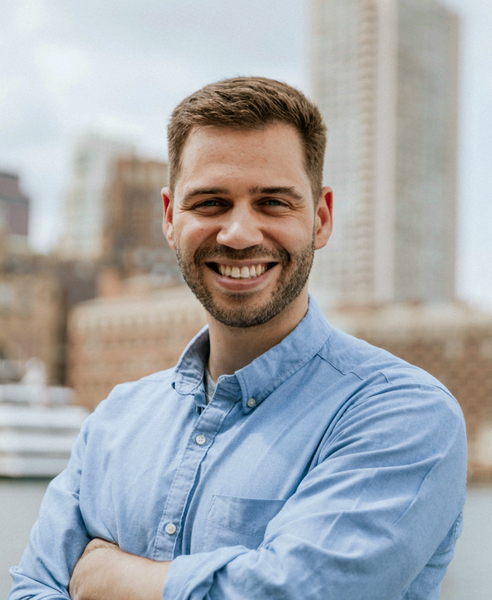 A man in a blue dress shirt with arms folded stands in front of a distant urban skyline.